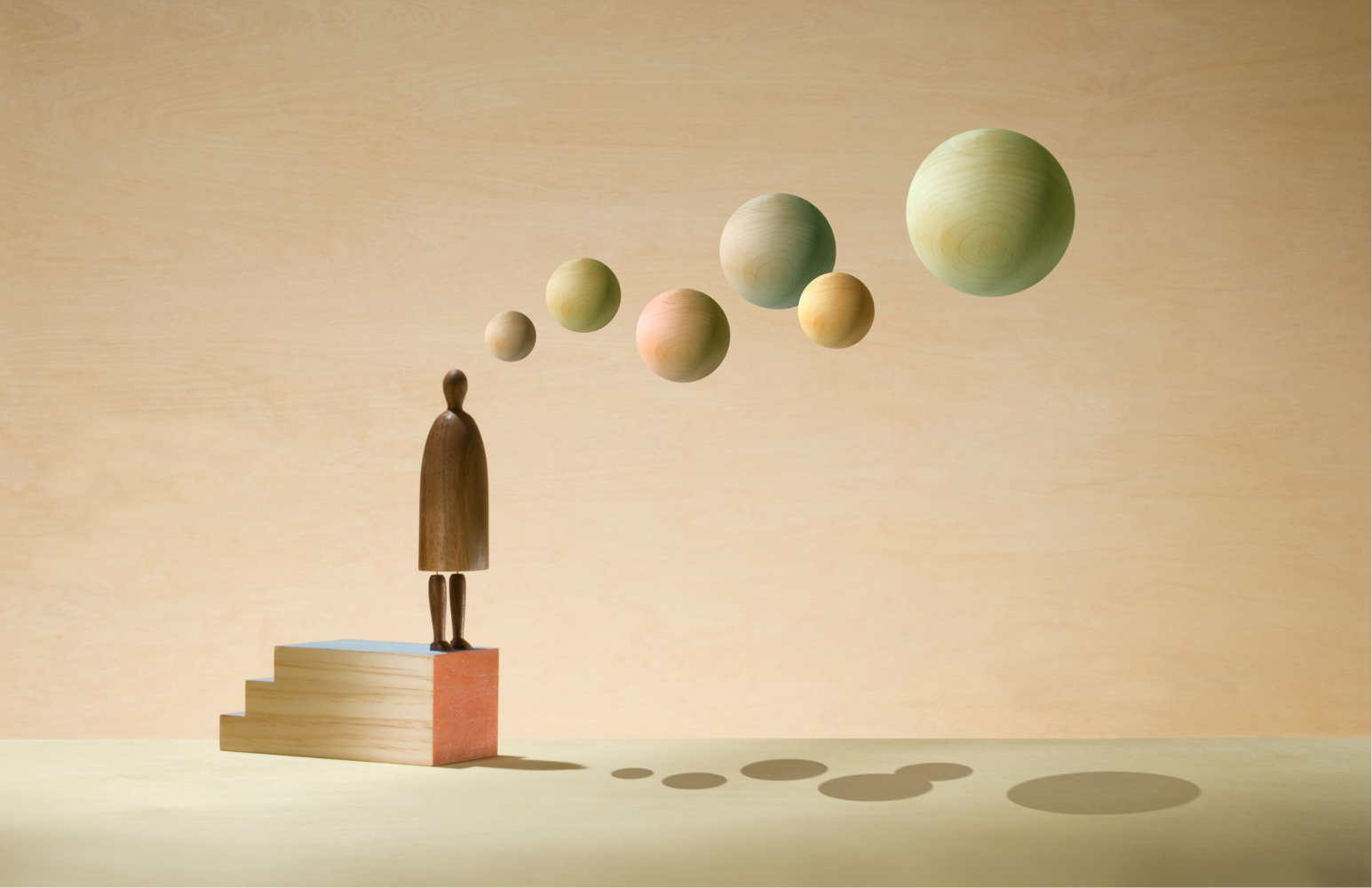 Wooden figurine stands on stacked blocks, watching pastel spheres float in a line like planets, casting shadows on the floor.